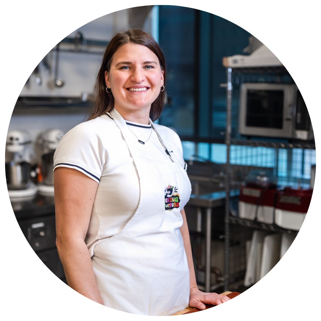 A woman stands smiling in a professional kitchen setting. She has straight brown hair, wears hoop earrings, and is dressed in a white t-shirt with black trim and a white apron that has a colorful logo reading "Cooking with Purpose." Behind her are kitchen appliances including stand mixers, a microwave, and various kitchen equipment organized on metal shelves. The image is cropped in a circular frame.
